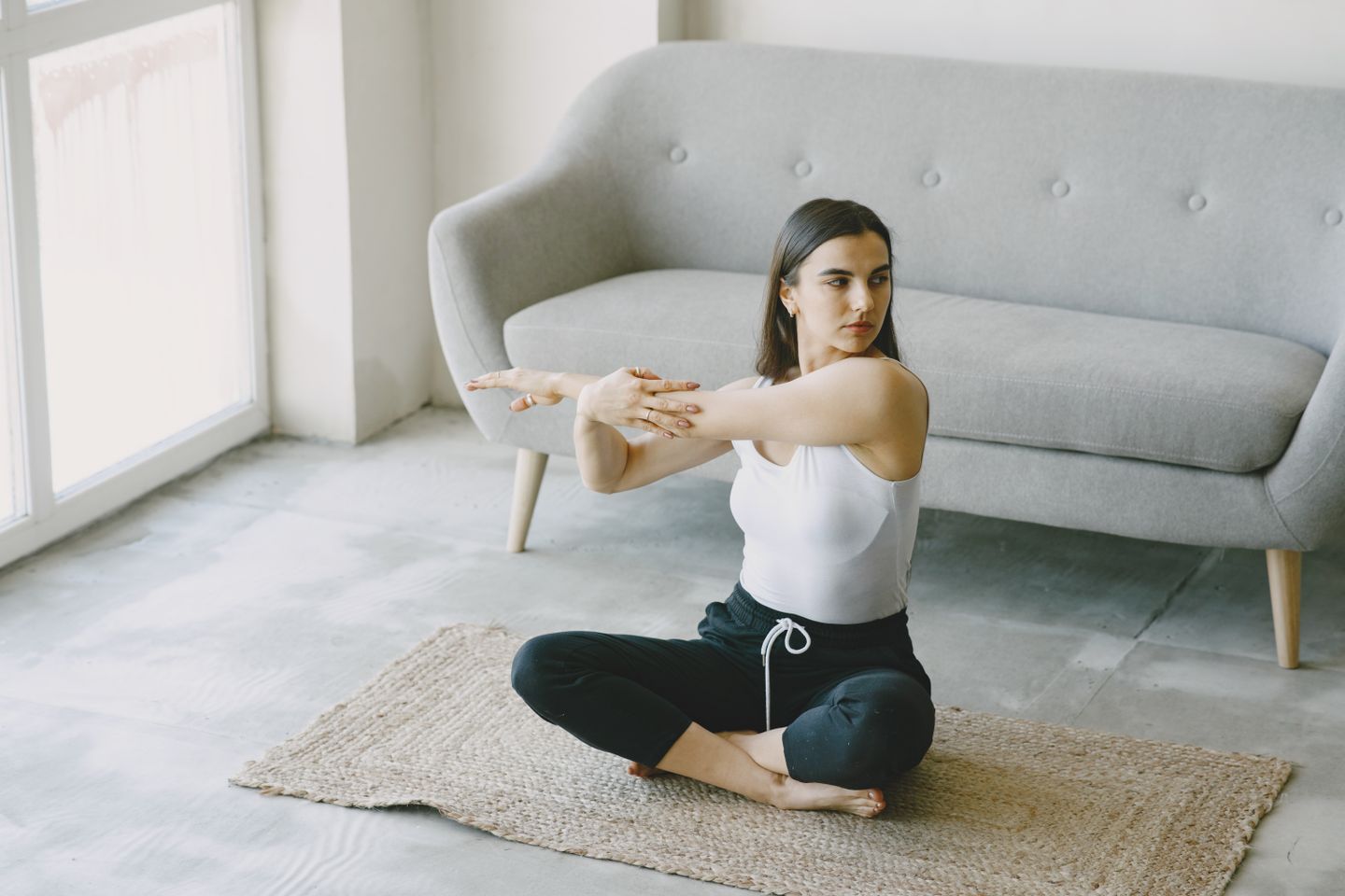 A woman in a tank top stretches while doing yoga indoors on a woven rug, near a sofa.